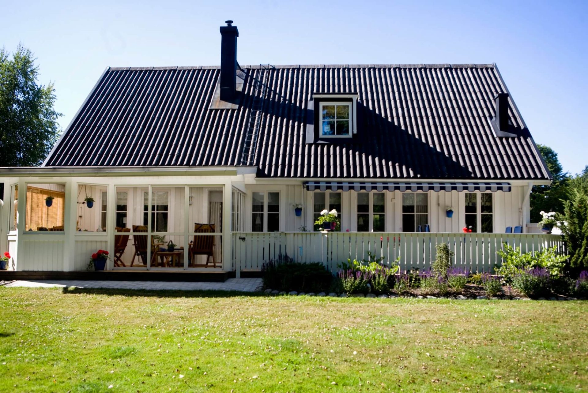 En vit trävilla under ett svart tak, kantad av en vid veranda, står prydligt i en trädgård rik på grönska